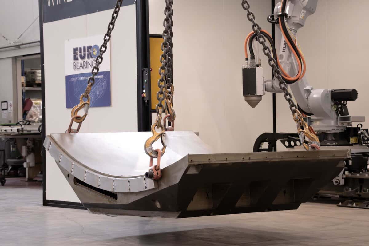 A large-scale industrial component manufactured using Meltio DED 3D Printing technology, suspended by chains at the Eurobearings facility in Cortemaggiore, Italy, with the KUKA robotic cell visible in the background