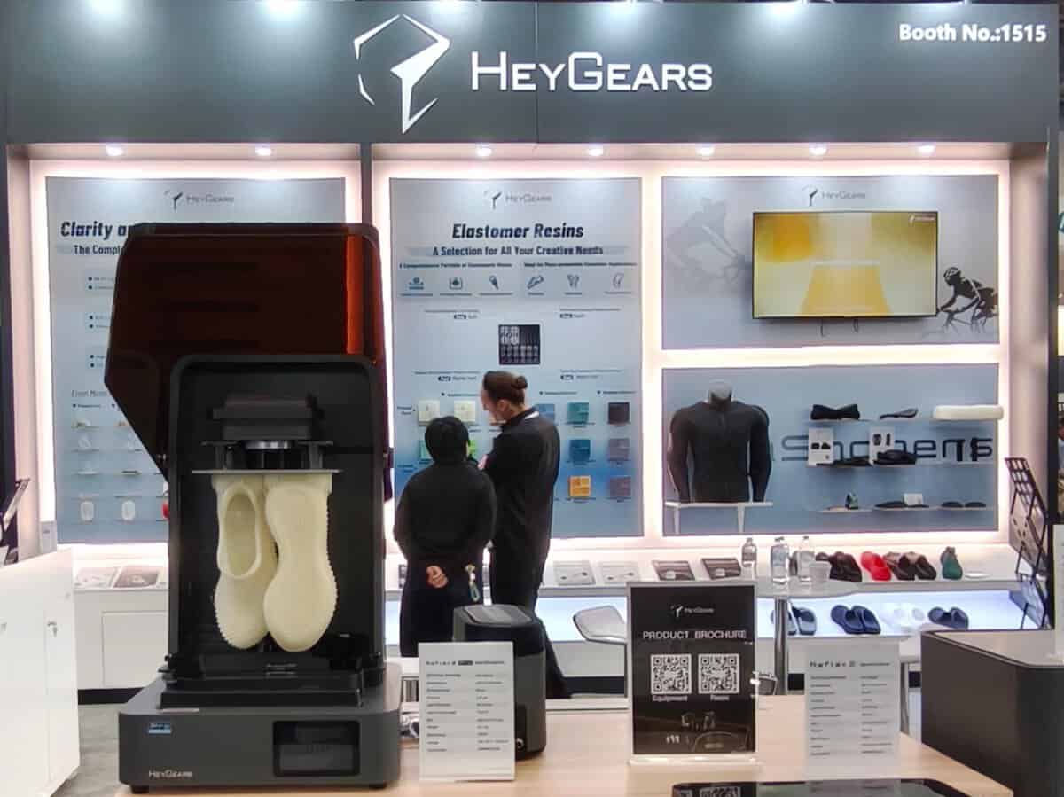 HeyGears' booth (No. 1515) at RAPID + TCT 2026 featuring a Reflex series 3D printer, elastomer resin samples, product brochures, and Transparent 3D Printing application parts
