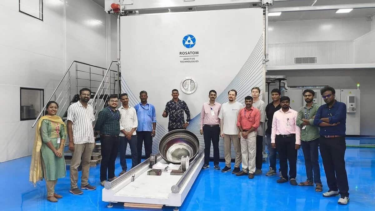 Rosatom Additive Technologies and ISRO engineers standing in front of the RusBeam 2800 electron-beam wire deposition system inside an industrial facility with blue flooring, with a large 3D printed metal component displayed on a platform in the foreground and Rosatom 3D Printer India branding visible on the machine
