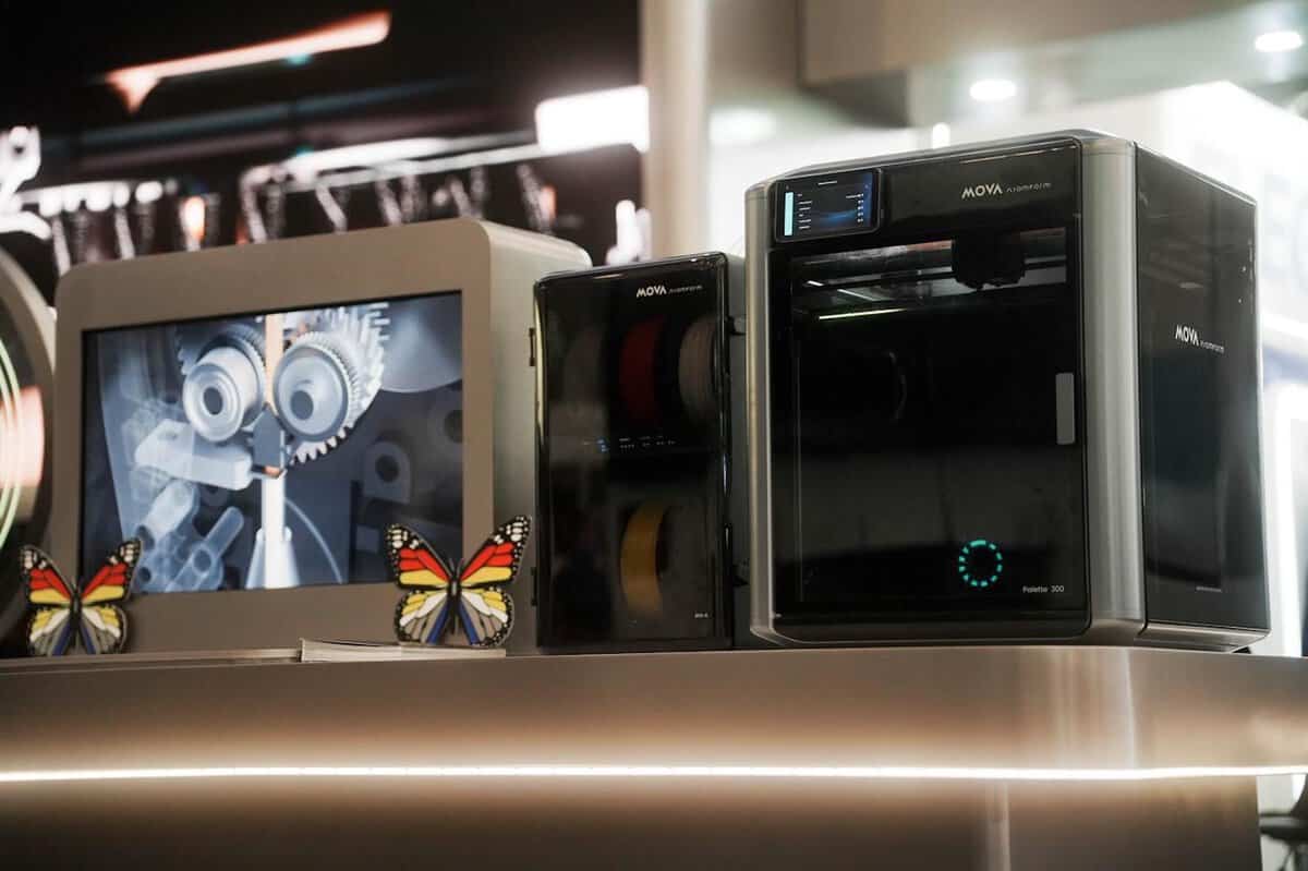 The MOVA AtomForm Palette 300 3D printer displayed alongside multi-colour 3D printed butterfly samples and a material storage unit at the RAPID + TCT 2026 exhibition booth