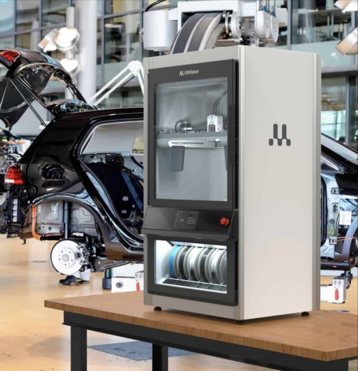 The UltiMaker Factor 4 Plus 3D printer positioned on a workbench inside an automotive assembly facility with vehicle chassis visible in the background