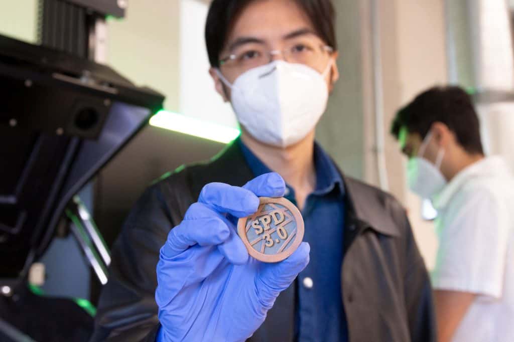 Close-up of Zhengtao Gan wearing blue nitrile gloves and holding a circular multi-metal additive manufacturing part with visible copper and steel sections marked "SPD 3.0," produced using the Aconity MIDI+ system at ASU's Reactive Materials 3D Printing Lab