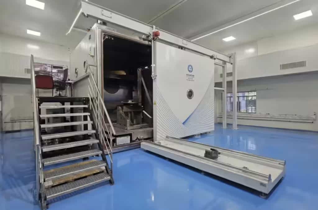 The Rosatom 3D Printer India RusBeam 2800 industrial system with its large sliding vacuum chamber door open, revealing the internal build chamber and electron-beam assembly, a retractable build platform extended outward on rails, metal access staircase to the left, and Rosatom Additive Technologies branding on the door panel, installed in a clean industrial facility with blue epoxy flooring