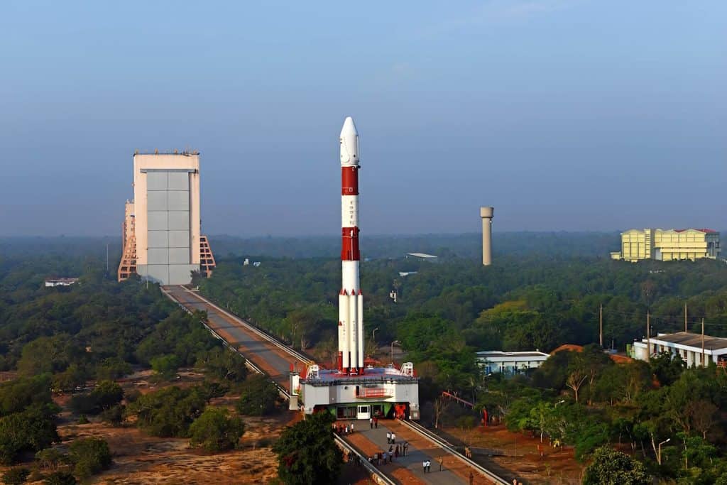 Aerial view of the Satish Dhawan Space Centre (SDSC) at Sriharikota relevant to 3D printing in Indian aerospace, showing a PSLV rocket standing vertically on the launch pad in the centre of the frame, the Vehicle Assembly Building visible to the left connected by a rail track, surrounding dense green vegetation, additional support buildings and a water tower to the right, under a hazy blue sky