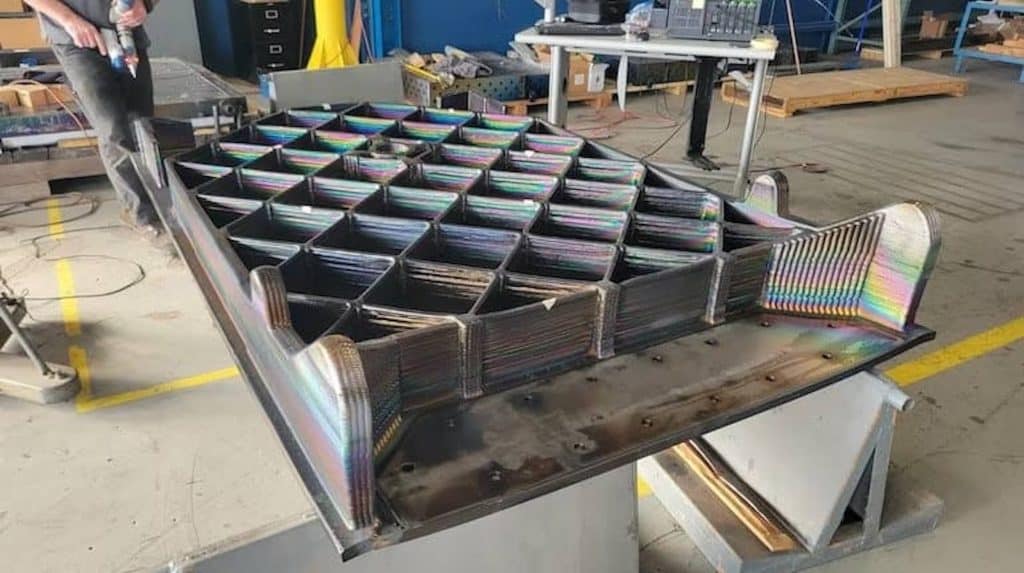 Close-up of a large titanium grid fin structure produced via electron-beam additive manufacturing, showing a cross-hatched lattice pattern with iridescent rainbow heat tinting on the layered metal deposition surfaces, positioned on a workbench in a workshop setting