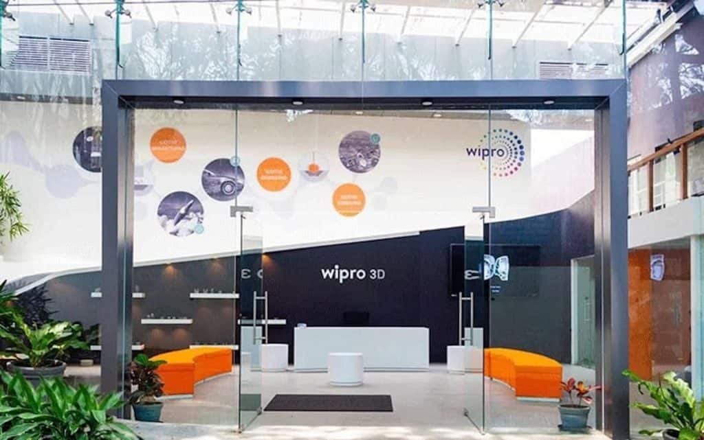Exterior entrance of the Wipro 3D Metal AM Solutions Centre in Bengaluru relevant to 3D printing in Indian aerospace, showing a modern glass-fronted reception area framed by a dark metal entryway with the Wipro 3D logo above, orange curved seating flanking the entrance, circular orange and grey display graphics on the left wall showcasing applications, metal AM component display shelves visible inside, and lush green plants surrounding the walkway